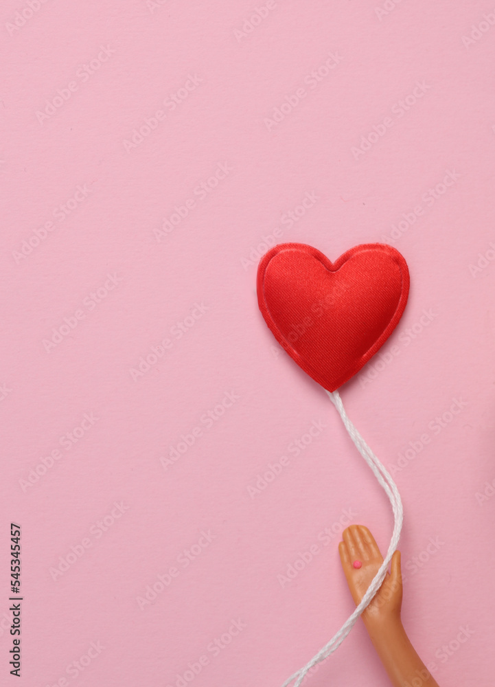 The doll's hand holds a heart-shaped air balloon on a pink background. Love concept