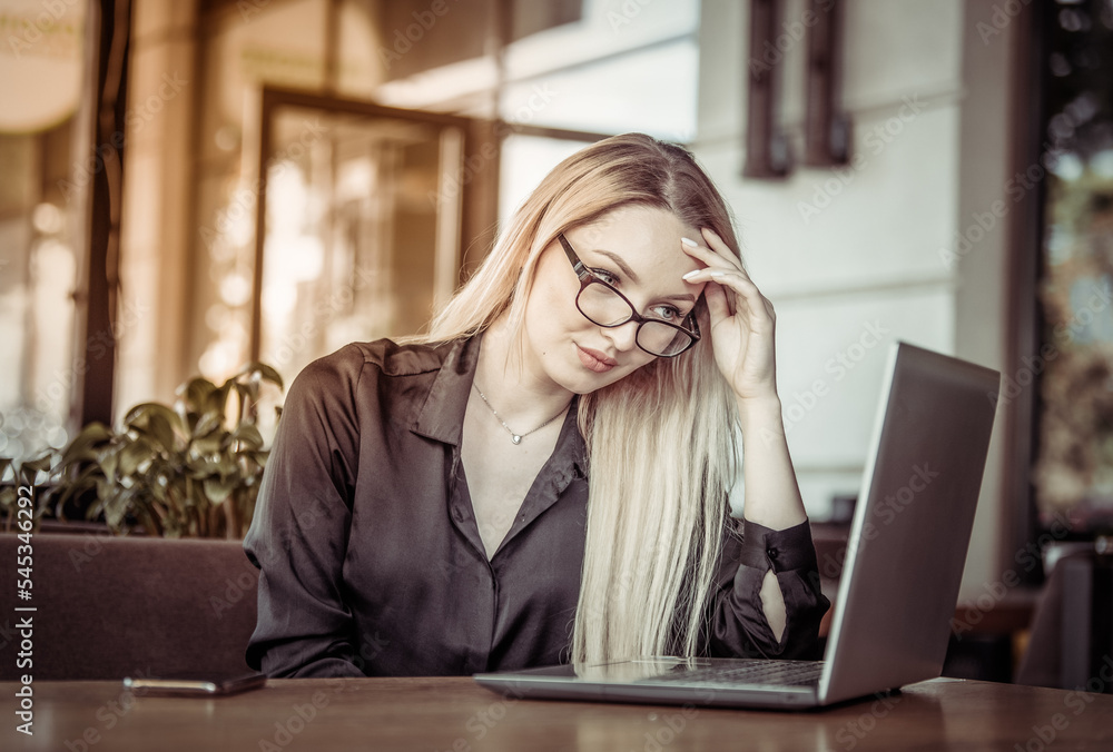 Tired blonde business woman looks into the laptop screen while sitting at a table in an outdoor cafe. Freelance, remote work Tired blonde business woman looks into the laptop screen while sitting at a table in an outdoor cafe. Freelance, remote work