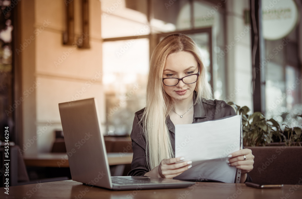 Blond Caucasian business woman examines sheets of documents and sits at a table with a laptop in an outdoor cafe. Modern business woman. Lifestyle Blond Caucasian business woman examines sheets of documents and sits at a table with a laptop in an outdoor cafe. Modern business woman. Lifestyle