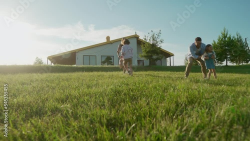 Happy positive parents playing with children outdoors near new house running and having fun. Family day activity. Kids play with mom and dad on grass on summer sunny day. Childhood, happiness