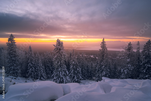 Winter sunrise on the top of the mountain with the sun shines through the clouds above snow-covered trees. © Sergey Egorov