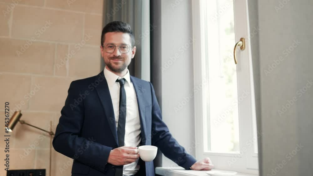 Handsome cheerful male in suit drinking coffee and looking at camera smiling in good mood. Portrait of young Caucasian happy businessman with cup of coffee looking at window indoor. Business style Handsome cheerful male in suit drinking coffee and looking at camera smiling in good mood. Portrait of young Caucasian happy businessman with cup of coffee looking at window indoor. Business style