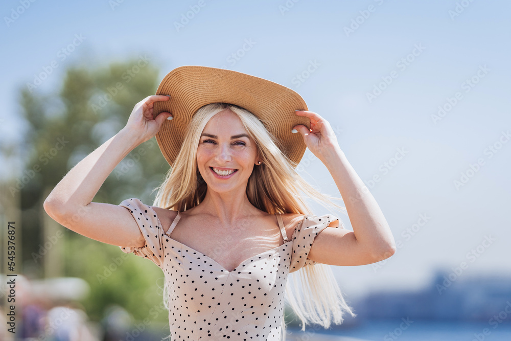 Cheerful blonde young Scandinavian woman in beige dress, straw hat toothy smiling against green trees and sea on background. Playful Italian girl traveling on vacations having fun. Leisure activities.