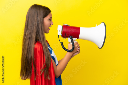 Little caucasian superhero girl isolated on yellow background shouting through a megaphone