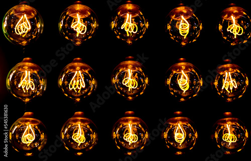 Isolated group of 15 yellow gold luminous retro classical spherical light bulbs in 3 rows with glowing filaments and black background 