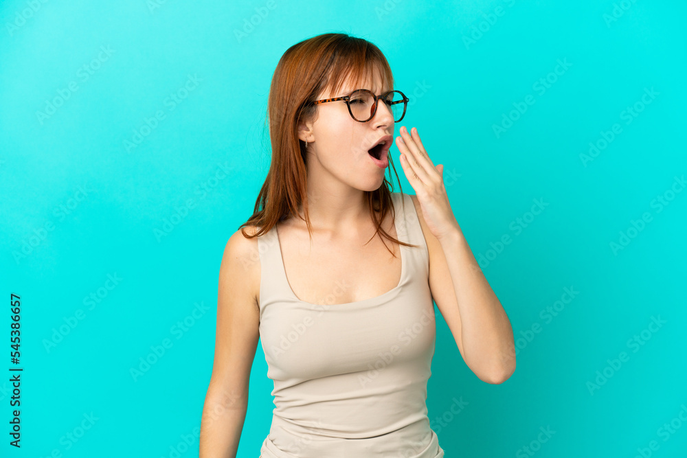 Redhead girl isolated on blue background yawning and covering wide open mouth with hand Redhead girl isolated on blue background yawning and covering wide open mouth with hand