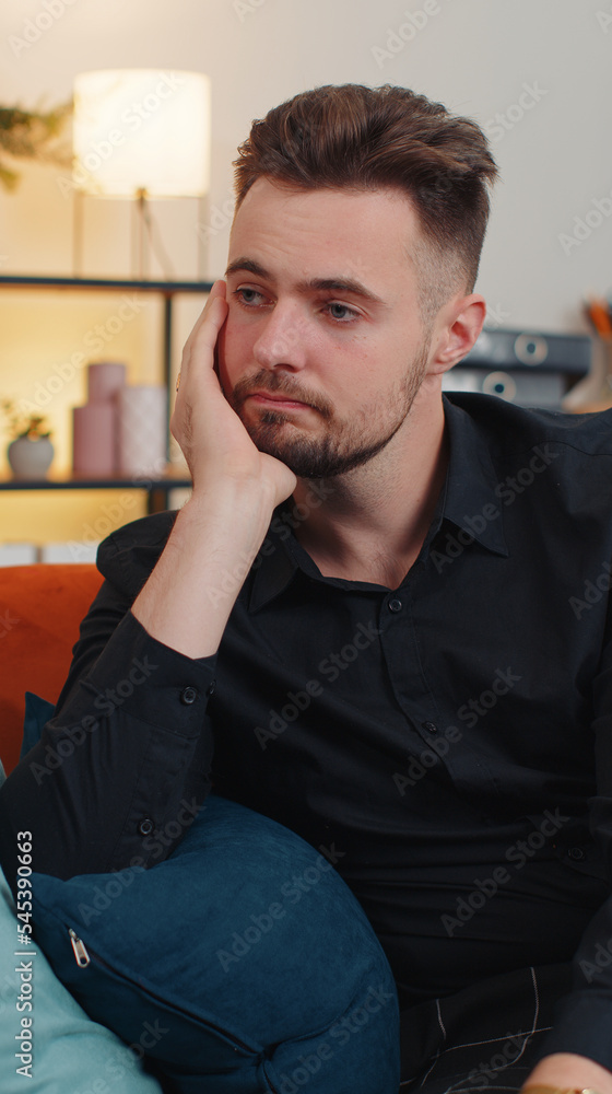 Fototapeta premium Sad man sitting at home looks pensive thinks over life concerns or unrequited love, suffers from unfair situation. Young guy problem, break up, depressed feeling bad annoyed, burnout. Vertical view