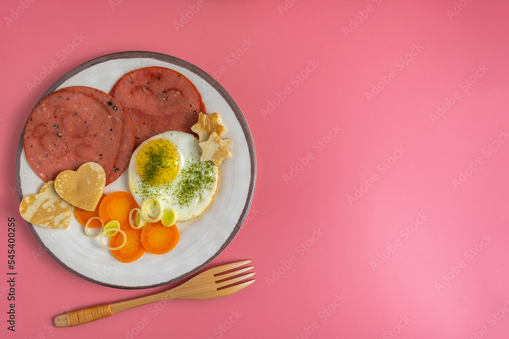 Breakfast plate with fresh carrots, miniature pancakes in the form of heart, sunny side up egg, roasted Bologna sausage over pink background. Free space for text. Breakfast plate with fresh carrots, miniature pancakes in the form of heart, sunny side up egg, roasted Bologna sausage over pink background. Free space for text.