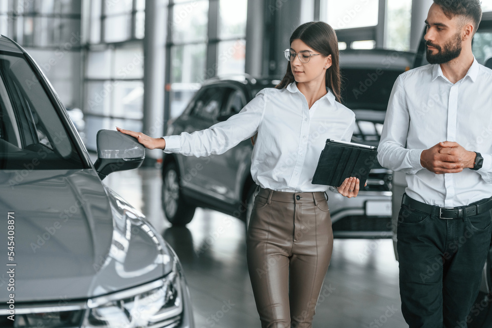 Front view. Man with woman in white clothes are in the car dealership together Front view. Man with woman in white clothes are in the car dealership together