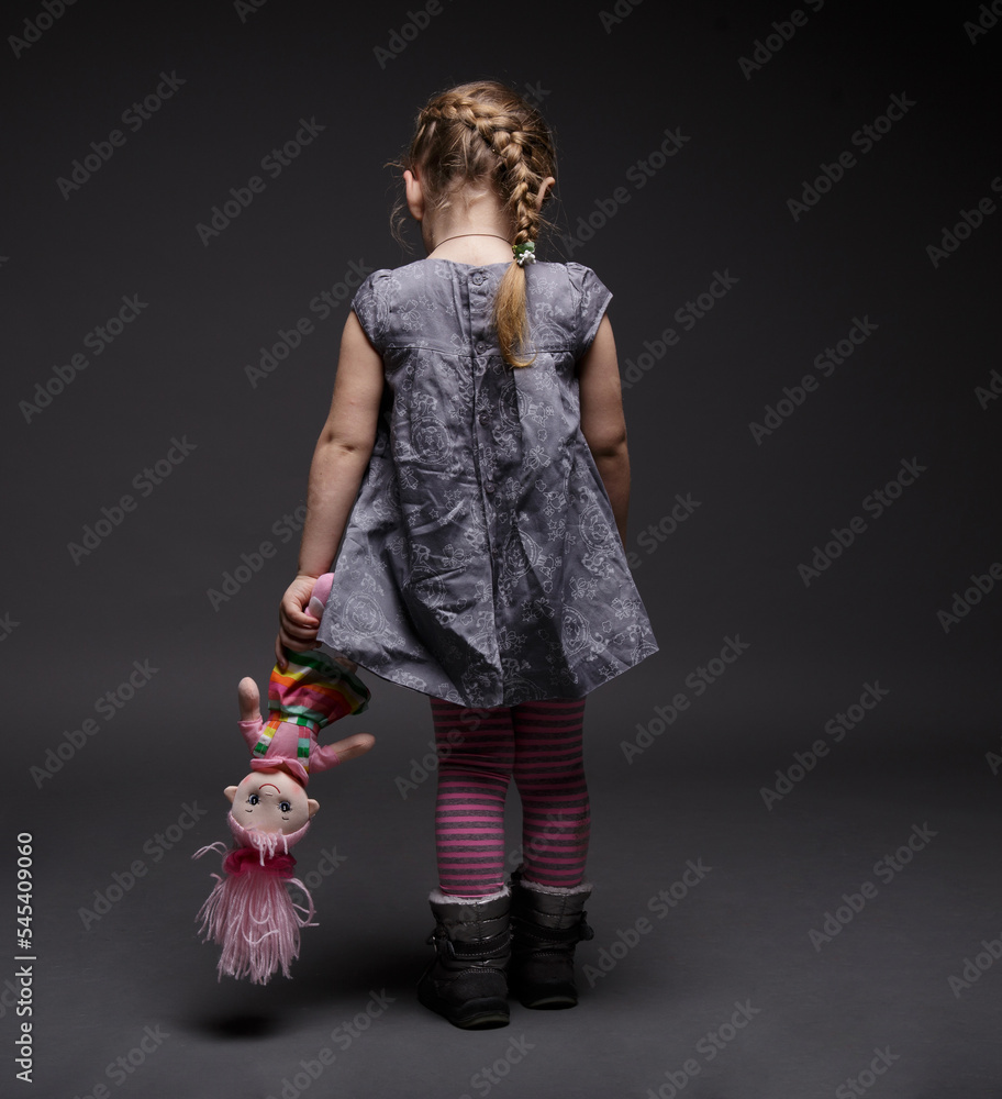 Rear image of a little sad girl with doll toddler offended by someone being in bad mood over dark grey background. Stock Photo Adobe Stock