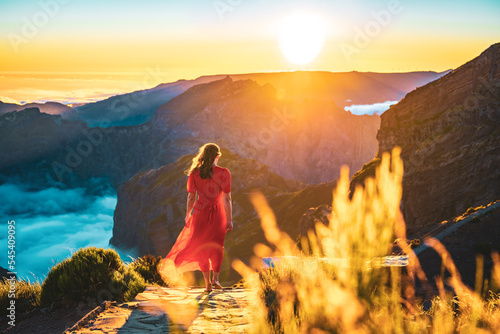 Beautiful woman in red dress walking barefoot on a very scenic hiking trail in the evening sun on Pico do Ariero. Verade do Pico Ruivo, Madeira Island, Portugal, Europe.