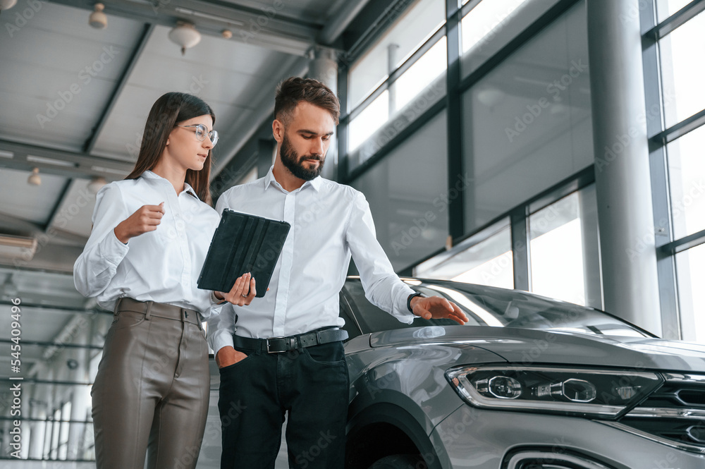 Black tablet is in the hands. Reading information. Man with woman in white clothes are in the car dealership together Black tablet is in the hands. Reading information. Man with woman in white clothes are in the car dealership together