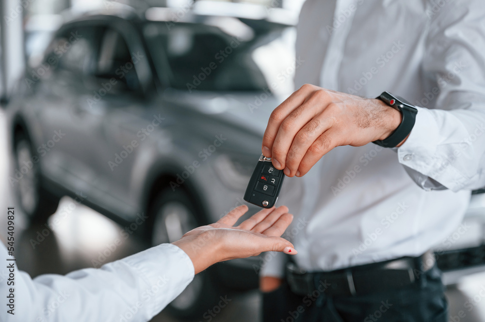 Keys of new automobile. Close up view. Man with woman in white clothes are in the car dealership together Keys of new automobile. Close up view. Man with woman in white clothes are in the car dealership together