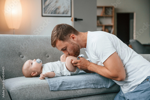 Baby is laying down on the sofa. Father with toddler is at home, taking care of his son