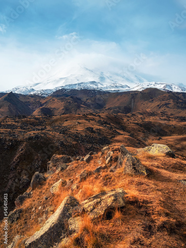 Bright white snowy peak of Mount Elbrus above the autumn rocky plateau. Autumn Elbrus. Autumn in the Caucasus mountains. White snow big mountains. Snow peaks. Vertical view.