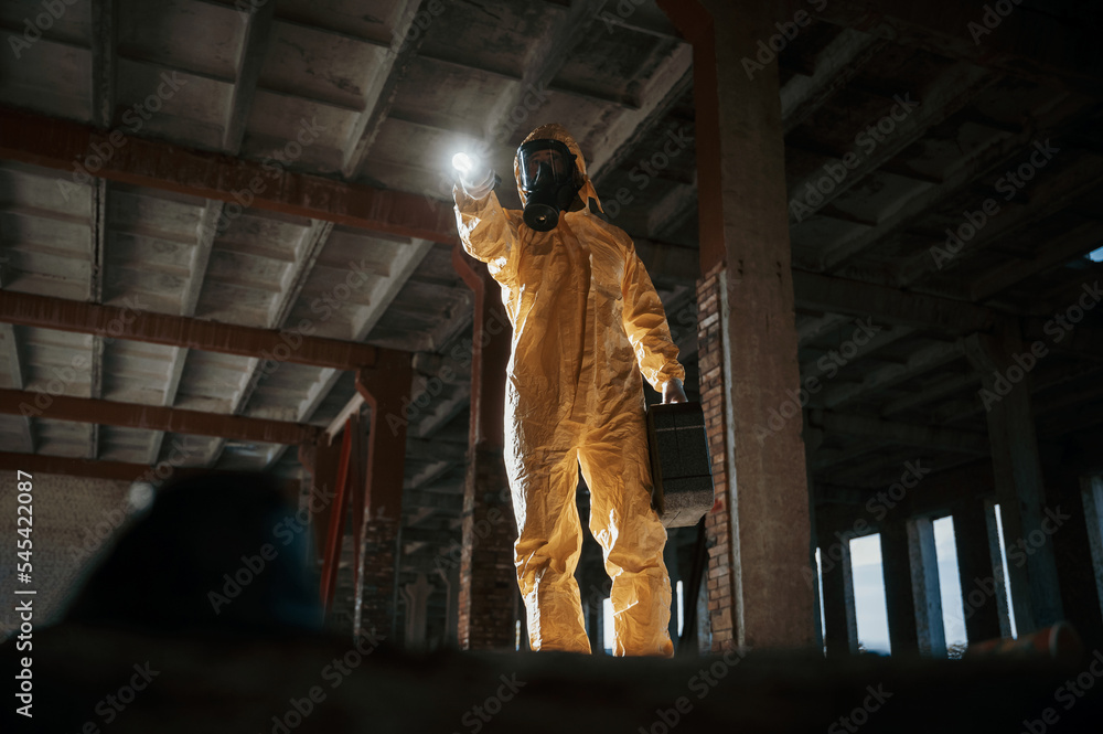 Fototapeta premium Using the flashlight. Man dressed in chemical protection suit in the ruins of the post apocalyptic building