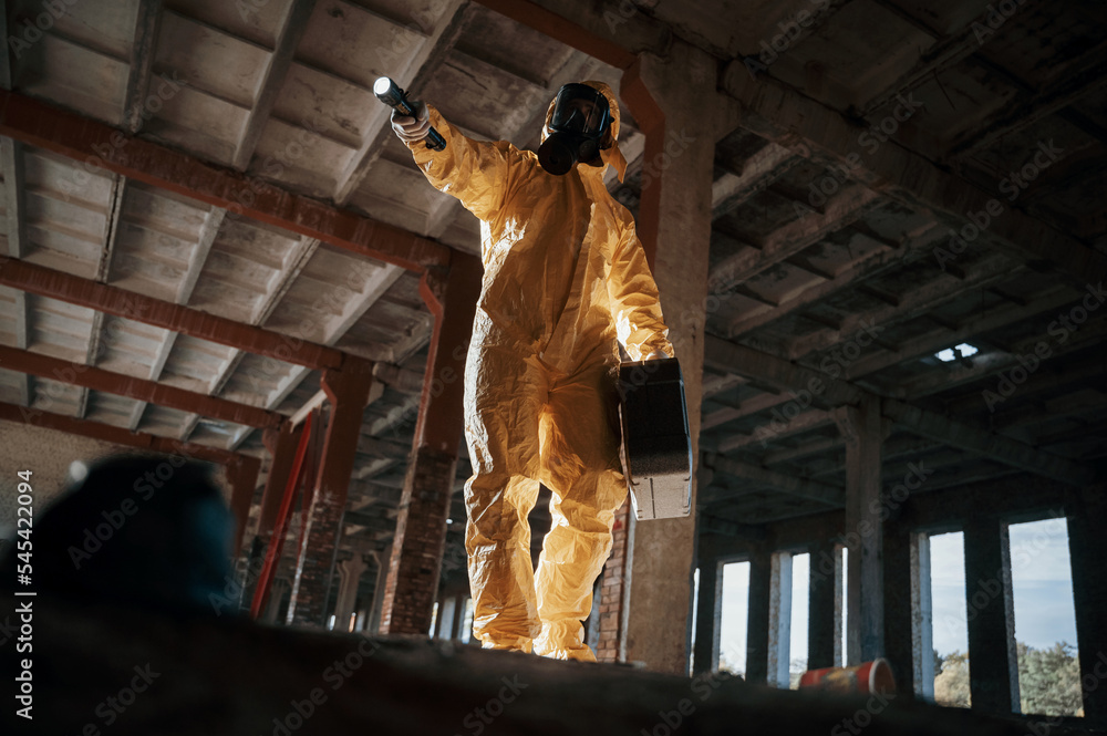 Using the flashlight. Man dressed in chemical protection suit in the ruins of the post apocalyptic building