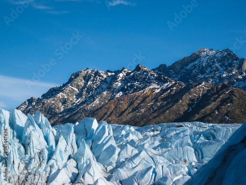 Glacial seracs of Matanuska Glacier in foreground, snow dusted mountains in background, and bluebird sky above
