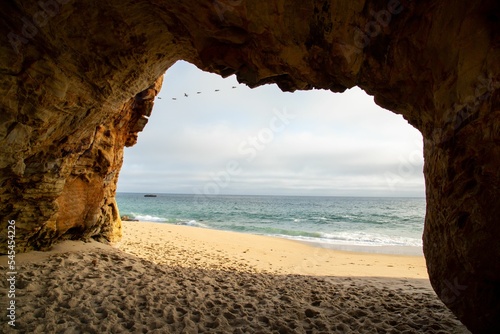 Fototapeta Closeup of a cave with a limestone archway, sandy beach, sea, and cloudy sky bac