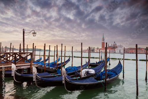 A row of gondolas parked beside the Riva degli Schiavoni in Venice, Italy. View of the Venetian Lagoon. The Church of San Giorgio Maggiore on island of the Venice, Italy, Europe © Andrey