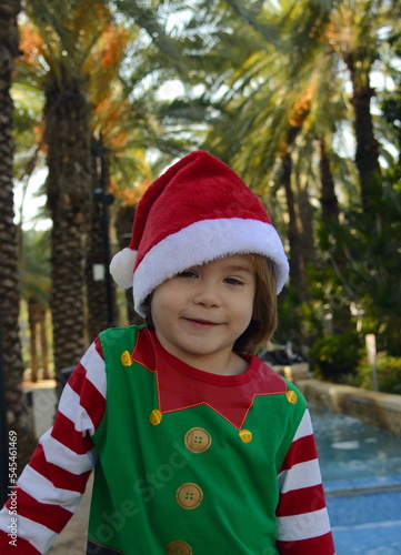 The child is waiting for a miracle, surprise, gift, holiday. A boy in a Santa Claus hat and an elf costume holds a finger to his lips: "silence, secret, do not speak" In the background of a palm tree,