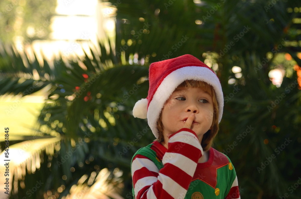 The child is waiting for a miracle, surprise, gift, holiday. A boy in a Santa Claus hat and an elf costume holds a finger to his lips: The child is waiting for a miracle, surprise, gift, holiday. A boy in a Santa Claus hat and an elf costume holds a finger to his lips: