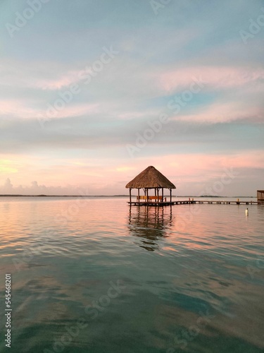 View of a sunset from lake Bacalar with a red and blue skyline and woody bridge, a roof in the water © Kevin281/Wirestock Creators