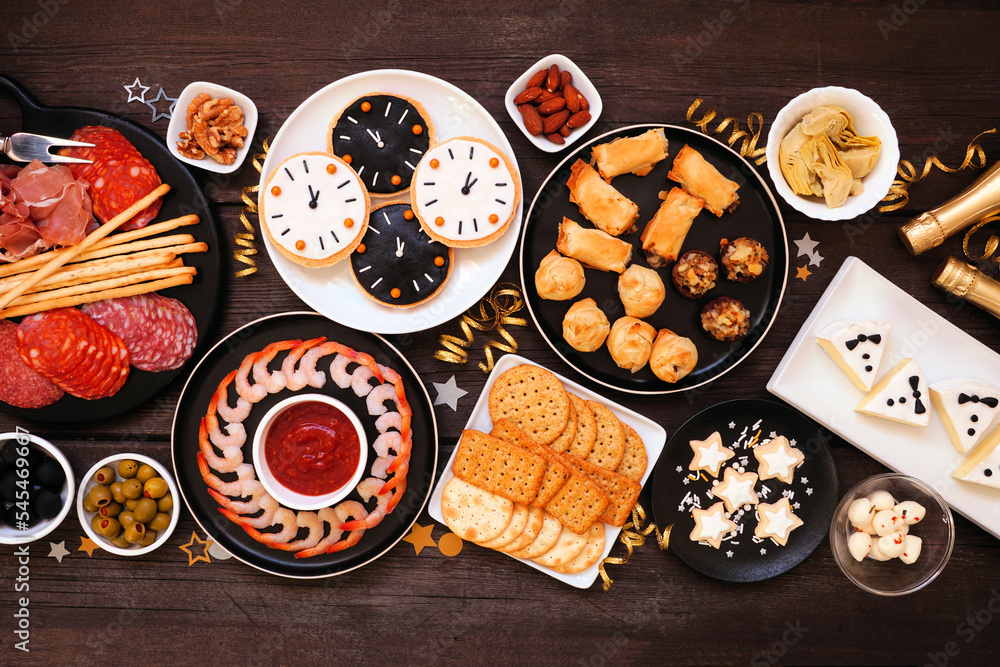 New Years Eve theme appetizer table scene. Above view on a dark wood background. Charcuterie board, champagne, clock cookies, tuxedo brie cheese and an assortment of party food. - obrazy, fototapety, plakaty 