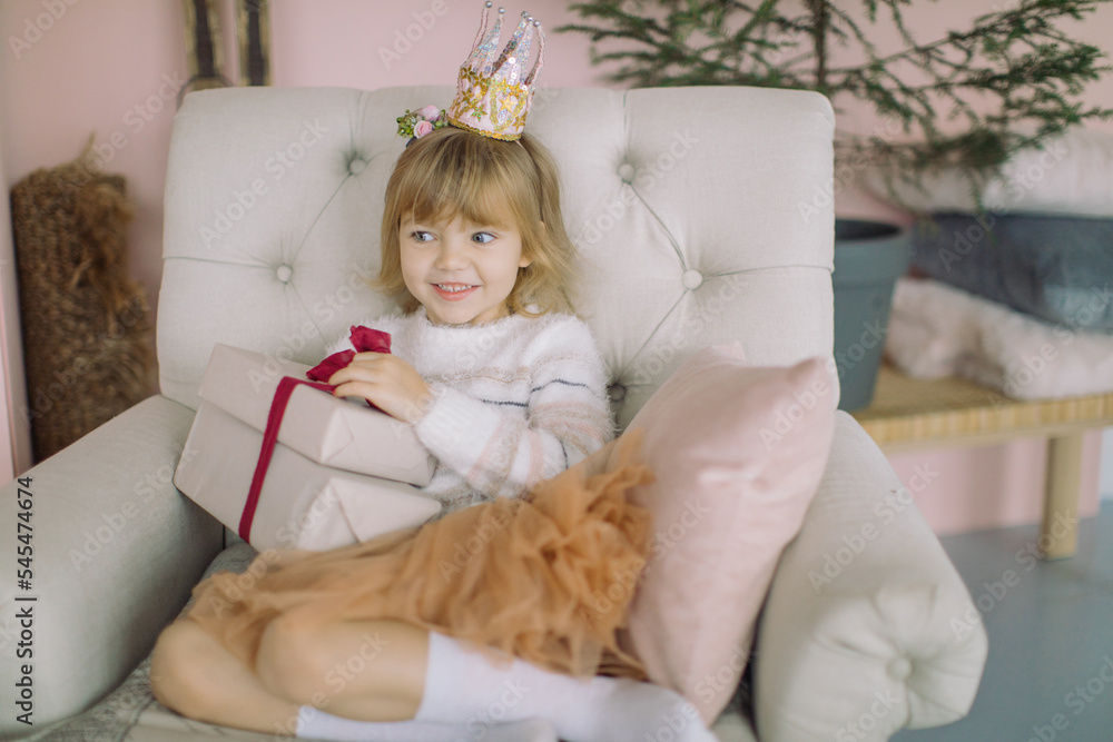Fototapeta premium Happy pretty girl in princess crown holding Christmas present in stylish home interior decorated for winter holidays. New Year, Christmas celebration concept.