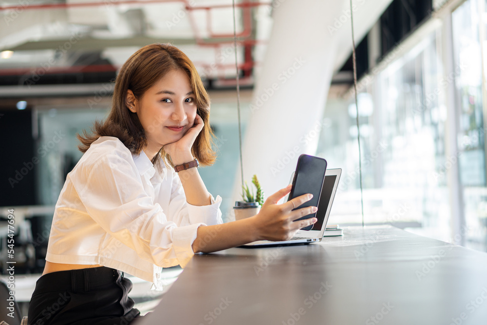 Attractive Asian woman talking on the phone and smiling while sitting at her office with laptop in the office.