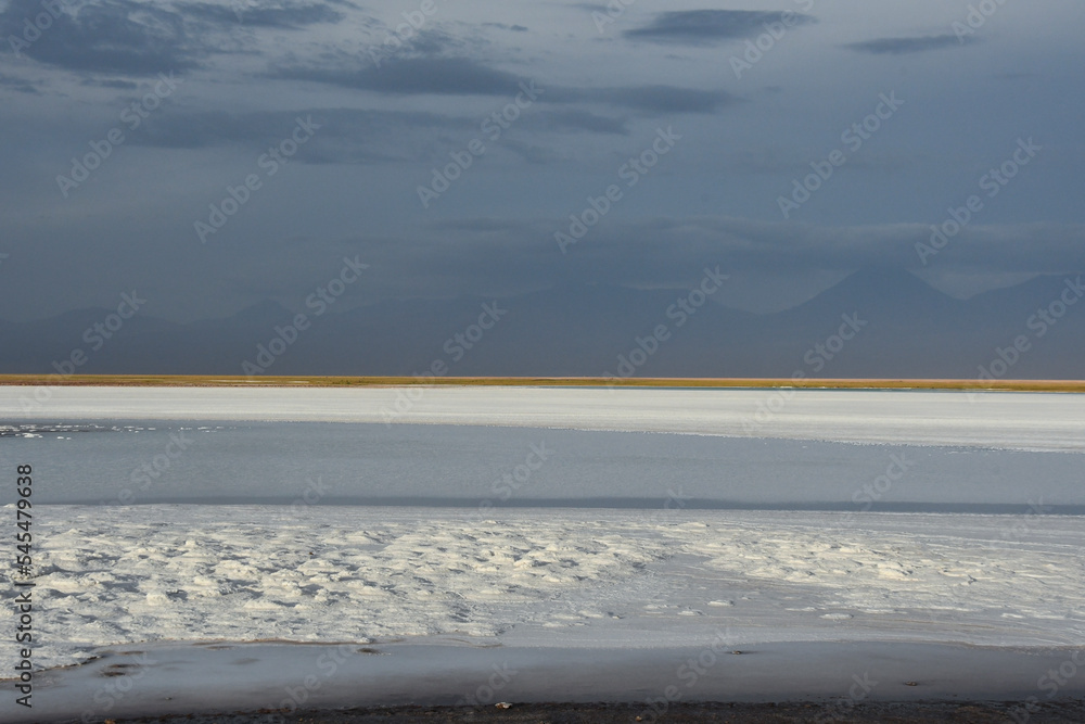 White salt flats in the Atacama desert at evening with mountains in the background and a yellow vegetation line on the horizon. Chile