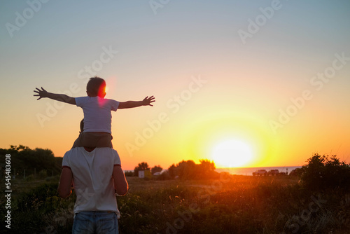 Dad carries a little son on his shoulders walking across the field against the backdrop of a sunset in the sky, back view