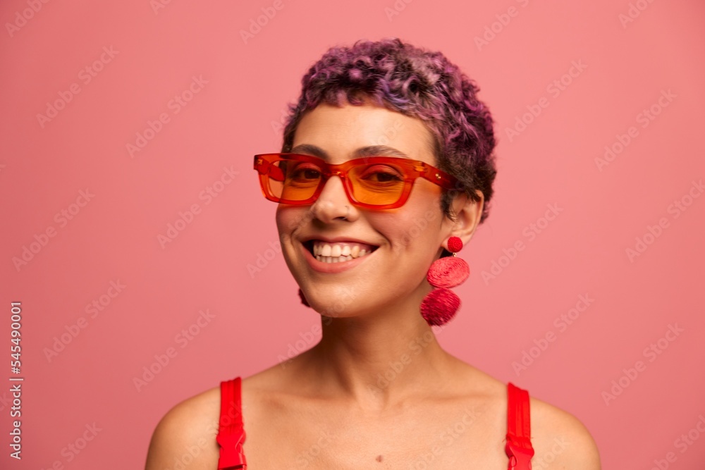 Fashion portrait of a woman with a short haircut in colored sunglasses with unusual accessories with earrings smiling on a pink bright background Fashion portrait of a woman with a short haircut in colored sunglasses with unusual accessories with earrings smiling on a pink bright background
