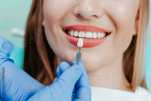 Smiling female mouth with natural white teeth in light blue background in dental clinic. Hands doctor dentist with teeth color palette next by face. Smile healthy teeth concept