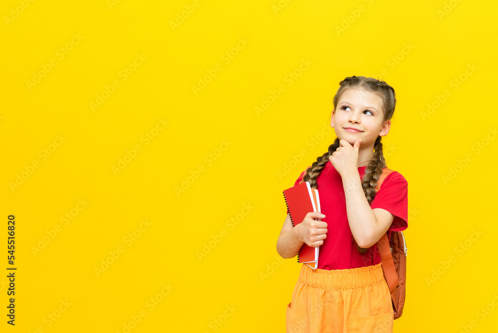 A schoolgirl with a satchel looks at your advertisement for educational courses for schoolchildren. A little girl is preparing for exams on a yellow isolated background. Copy space. A schoolgirl with a satchel looks at your advertisement for educational courses for schoolchildren. A little girl is preparing for exams on a yellow isolated background. Copy space.