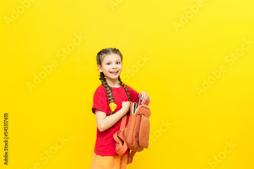 A schoolgirl is preparing a satchel for school. A little girl collects notebooks and textbooks for the school day on a yellow isolated background. Preparation for exams.