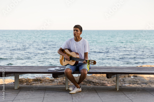 A young stylish guy plays the guitar on the embankment by the sea, the concept of music and relaxation © Enigma