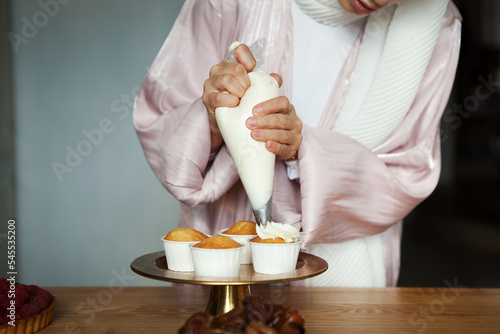 Woman cooks cupcakes, squeezes cream on cakes. Close-up of hands Muslim pastry chef girl preparing dessert