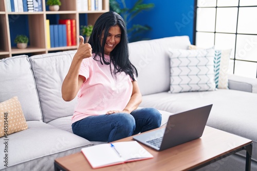Mature hispanic woman using laptop at home smiling happy and positive, thumb up doing excellent and approval sign