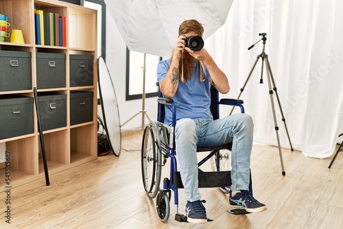Young redhead man photographer sitting on wheelchair using professional camera at photograpy studio photo