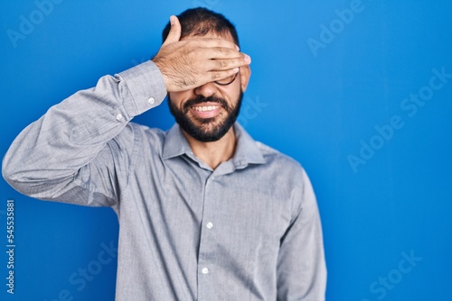 Middle east man with beard standing over blue background smiling and laughing with hand on face covering eyes for surprise. blind concept. © Krakenimages.com
