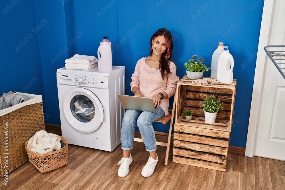Young caucasian woman using laptop waiting for washing machine at laundry room Young caucasian woman using laptop waiting for washing machine at laundry room