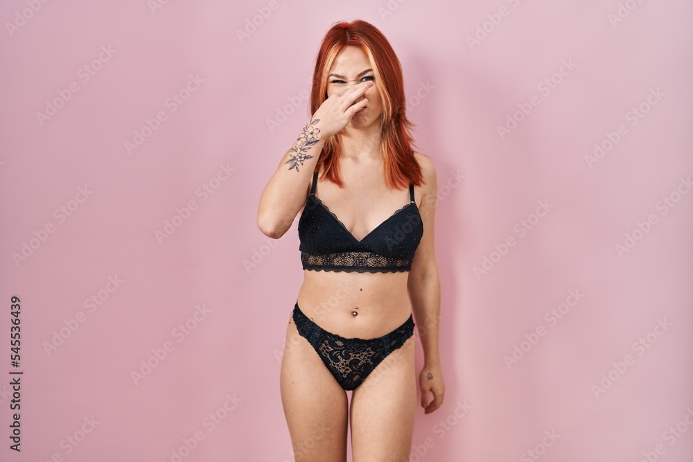 Naklejka premium Young caucasian woman wearing lingerie over pink background smelling something stinky and disgusting, intolerable smell, holding breath with fingers on nose. bad smell