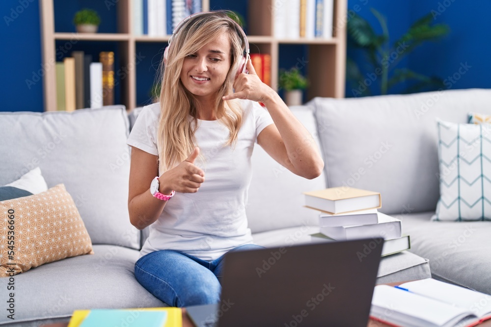 Young blonde woman studying using computer laptop at home smiling doing talking on the telephone gesture and pointing to you. call me. Young blonde woman studying using computer laptop at home smiling doing talking on the telephone gesture and pointing to you. call me.