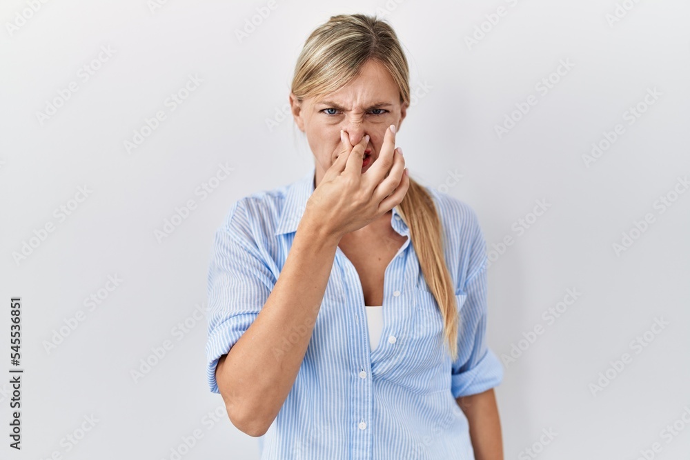 Beautiful blonde woman standing over white background smelling something stinky and disgusting, intolerable smell, holding breath with fingers on nose. bad smell Beautiful blonde woman standing over white background smelling something stinky and disgusting, intolerable smell, holding breath with fingers on nose. bad smell