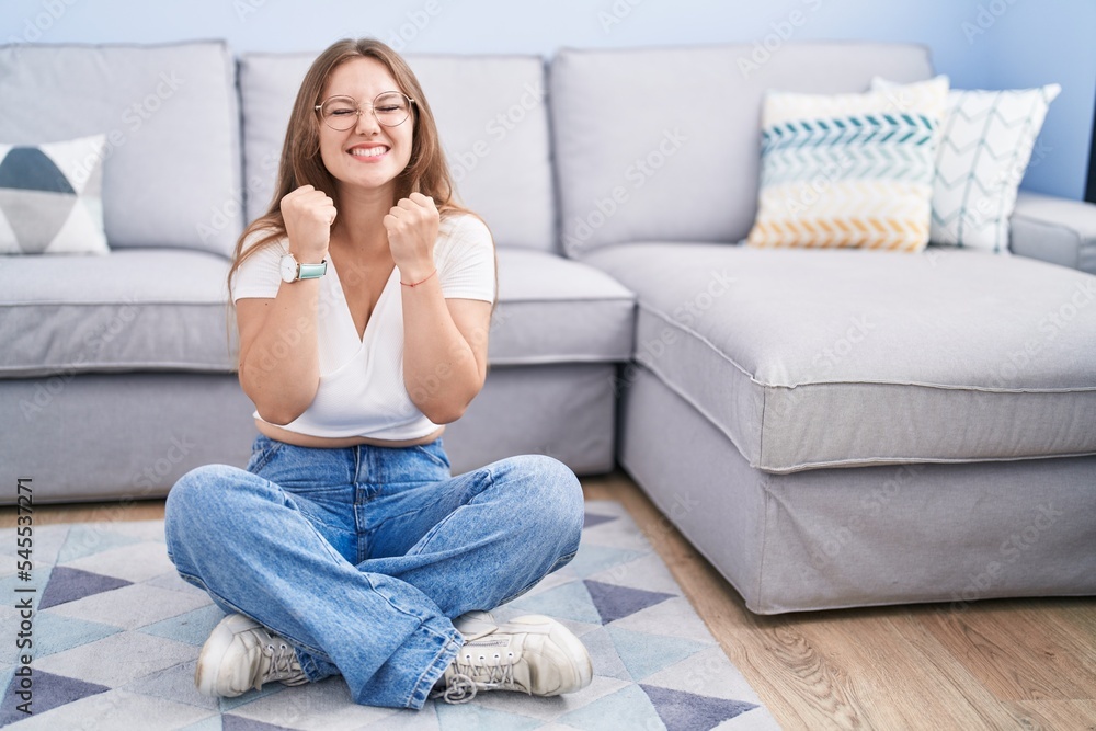 Young caucasian woman sitting on the floor at the living room excited for success with arms raised and eyes closed celebrating victory smiling. winner concept.