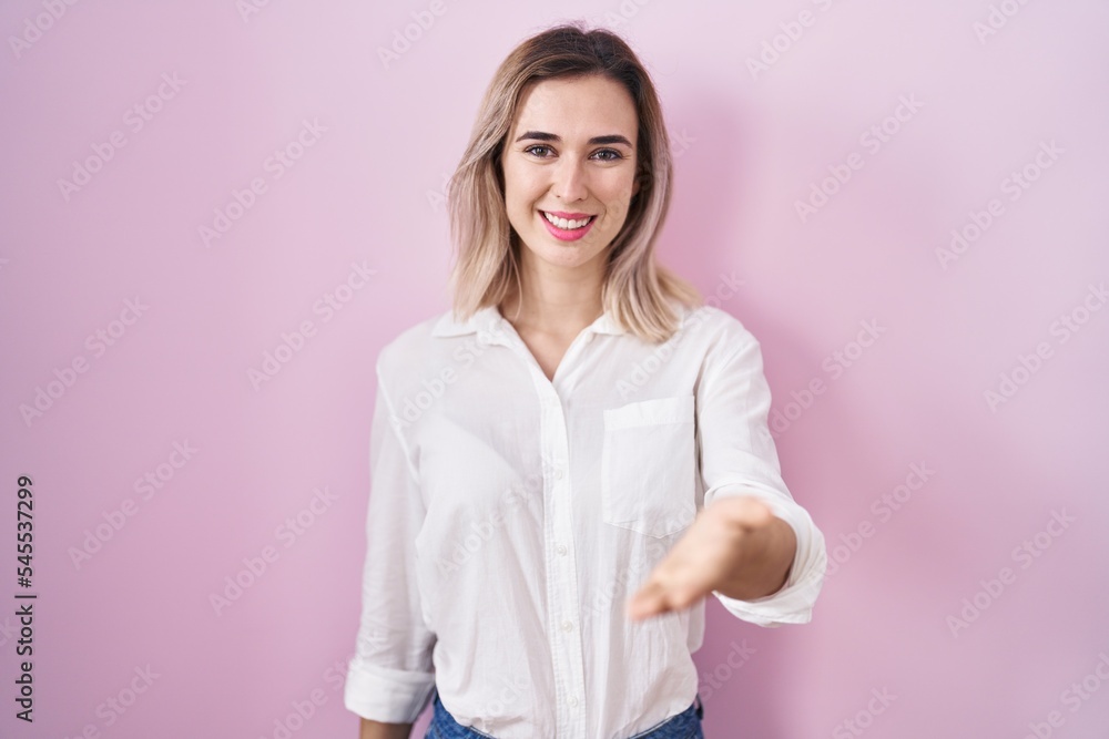 Young beautiful woman standing over pink background smiling cheerful offering palm hand giving assistance and acceptance. Young beautiful woman standing over pink background smiling cheerful offering palm hand giving assistance and acceptance.