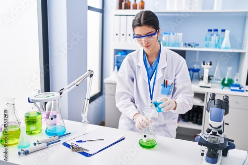 Young beautiful hispanic woman scientist pouring liquid on test tube at laboratory