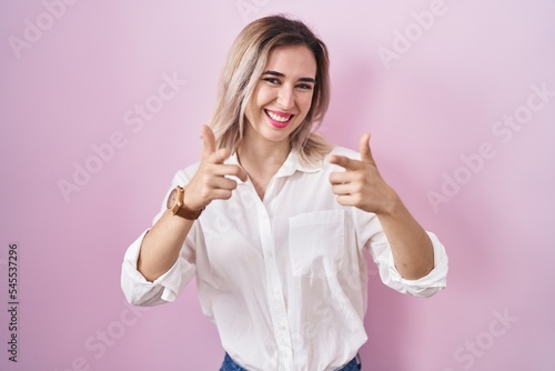 Young beautiful woman standing over pink background pointing fingers to camera with happy and funny face. good energy and vibes.