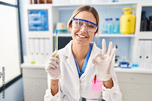Young blonde woman working at scientist laboratory holding sample doing ok sign with fingers, smiling friendly gesturing excellent symbol